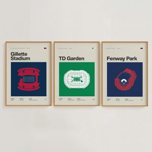 May include: Three framed art prints featuring stadium layouts. The prints showcase Gillette Stadium in red, TD Garden in green, and Fenway Park in red, each set against a contrasting background. The prints are in gold frames.