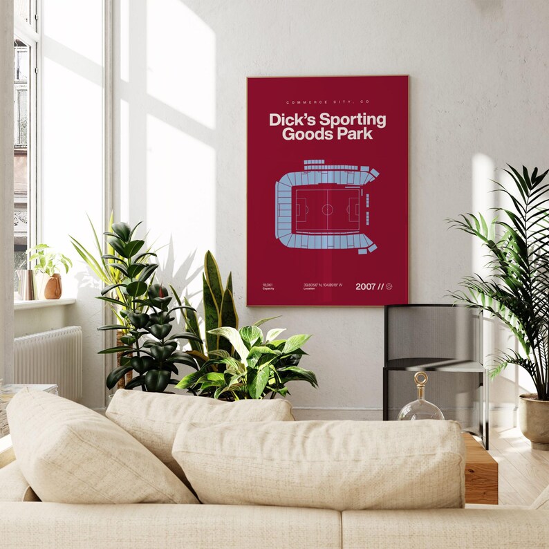 May include: A framed burgundy print of Dick's Sporting Goods Park stadium in Commerce City, CO. The stadium is depicted in light blue, with the text "Dick's Sporting Goods Park" in white. The print is displayed in a well-lit living room with plants and a beige sofa.