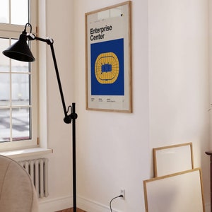 May include: A framed print of a blue and yellow stadium floor plan with the text "Enterprise Center" in white. The print is hanging on a white wall in a room with a black floor lamp and a white couch.