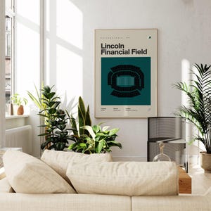 May include: A framed poster featuring the text "Lincoln Financial Field" with a teal and black stadium diagram. The poster is displayed in a well-lit living room with plants and a beige sofa.