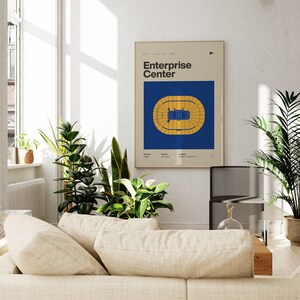 May include: A blue and yellow poster with the text "Enterprise Center" and a diagram of the arena floor plan. The poster is hanging on a white wall in a room with a couch and plants.