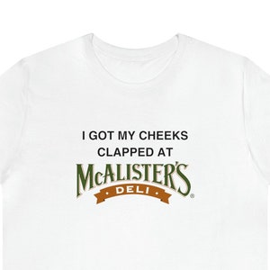 Op de afbeelding: Wit T-shirt met de tekst "I got my cheeks clapped at McAlister's Deli" in zwart gedrukt.