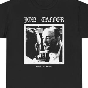 May include: Black t-shirt featuring a black and white image of two men in a heated discussion. The text "JON TAFFER" is above the image, and "SHUT IT DOWN" is below. The shirt is made of a soft material.