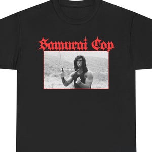 Puede incluir: Camiseta negra con el texto rojo "Samurai Cop" en letras góticas. Debajo, una imagen en blanco y negro de un hombre con una espada, enmarcada en rojo. La camiseta tiene cuello redondo.