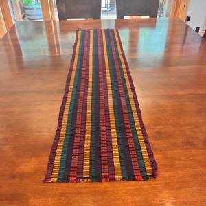 May include: A colorful table runner with stripes of red, yellow, green, and purple. The runner has a fringed edge and is made of a woven fabric.