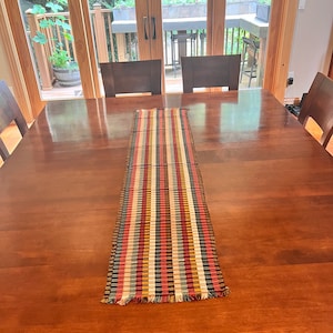 May include: A colorful striped table runner with fringed edges, laid out on a wooden table.