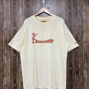 May include: A cream-coloured short-sleeve t-shirt featuring a brown graphic on the chest of a figure with an afro hairstyle pointing upward, accompanied by the text "KINGDIGGIN". A small brown circular logo is printed on the left sleeve.