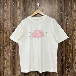 May include: A white short-sleeve t-shirt featuring a central graphic of a pink anatomical brain with various lines pointing to different sections, accompanied by small symbols and the text "Fig 1: The Human Brain" printed below.