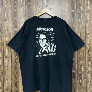 May include: A black short-sleeve t-shirt featuring a white graphic print on the back. The print displays the Microsoft logo above an illustration of a person pointing forward, with the text SEE YOU NEXT TUESDAY! printed in white capital letters.