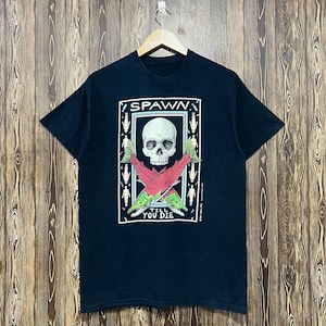May include: A black short-sleeved t-shirt featuring a graphic print of a human skull positioned above two crossed salmon. The text "SPAWN" appears above the skull and "TILL YOU DIE" appears below the fish, surrounded by small human figures.