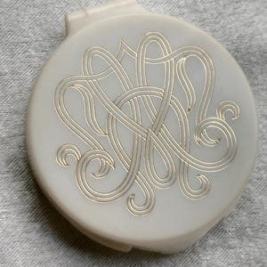 May include: A round, off-white compact with a gold-colored, intertwined monogram design. The compact is closed and has a small, hinged opening. The background is a textured, gray fabric.