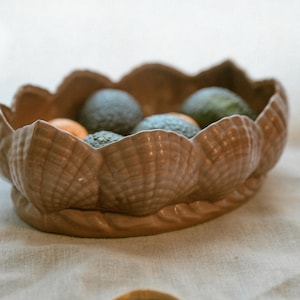 Puede incluir: Un cuenco de cerámica marrón con forma de concha, lleno de aguacates y pequeñas frutas naranjas. El cuenco está sobre una superficie de lino beige, con una pulsera de madera en primer plano. La textura y los colores naturales crean una estética rústica.