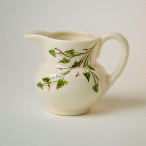 May include: A cream-colored ceramic creamer pitcher featuring a delicate hand-painted design of green ivy leaves and small pink berries. The pitcher has a rounded body, a curved handle, and a small pouring spout, showcasing a smooth finish.