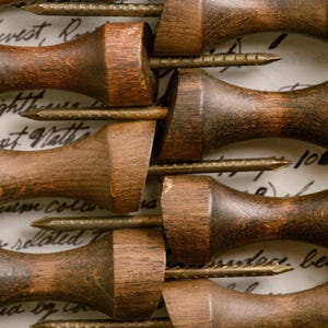 May include: Close-up of several antique wooden tools with brass nails. The tools have a dark brown finish and are arranged in parallel rows. The background features handwritten script.