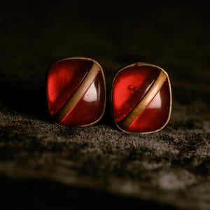 May include: A pair of square-shaped cufflinks featuring a translucent deep red resin or glass center. Each piece is bisected by a diagonal metallic bar and framed by a thin gold-toned metal border, creating a smooth, polished appearance.