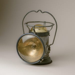 May include: A vintage metal lantern featuring a large, circular glass lens with a reflective gold-toned interior. The body is grey with a weathered finish, topped by a curved wire handle and a small, rounded yellow glass dome at the very top.