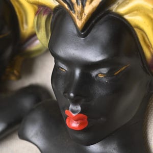 May include: Close-up of a black bust with red lips and gold accents. The figure has closed eyes and a decorative headdress with gold and yellow tones. The bust is set against a neutral background.