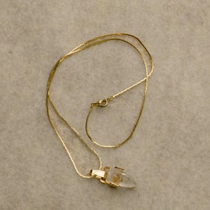 May include: A gold necklace featuring a clear crystal pendant. The chain is fine and the clasp is visible. The pendant is set in gold. The necklace is presented against a neutral backdrop.