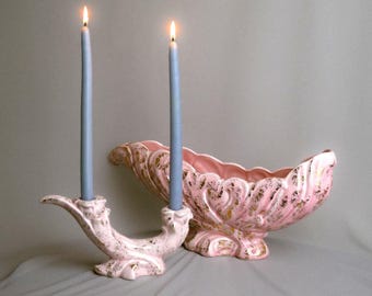 May include: A light pink ceramic candle holder with two lit blue taper candles and a matching pink ceramic bowl, both featuring ornate, scalloped edges and decorative gold-toned accents throughout the textured surfaces.