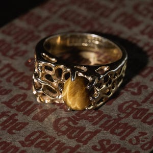 May include: A gold ring with an openwork design and a golden-brown gemstone. The ring is set on a brown surface with faded red text. The ring's design is intricate and detailed.