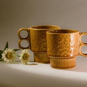 May include: Two amber-colored ceramic mugs featuring a raised floral pattern on the body and a ribbed base. Each mug has a distinctive handle composed of two stacked circular loops. Two white flowers with yellow centers are positioned nearby.