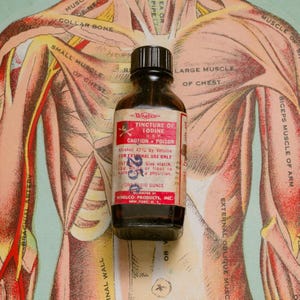 Könnte beinhalten: Eine Vintage-Braunglasflasche mit Wheeler's Tinktur aus Jod, mit schwarzem Verschluss und rot-weißem Etikett. Das Etikett warnt vor Gift und nur zur äußerlichen Anwendung. Die Flasche steht vor dem Hintergrund einer medizinischen Illustration.