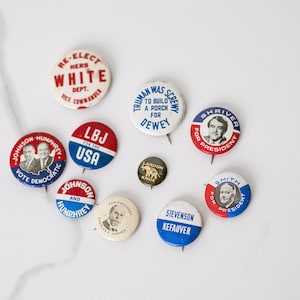 Könnte beinhalten: Eine Sammlung politischer Vintage-Ansteckbuttons. Die Buttons zeigen Text und Porträts von Kandidaten in den Farben Rot, Weiß und Blau. Einige Buttons tragen Aufschriften wie "Re-Elect White" und "LBJ for the USA".