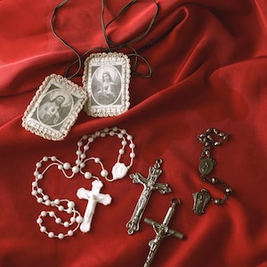 May include: Two rectangular scapulars with black cords, featuring black and white religious imagery and lace-like borders. A white beaded rosary with a white crucifix, a silver-toned metal crucifix, and a small dark metal rosary chain.