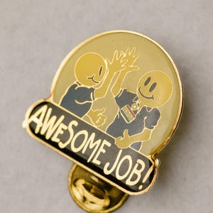 Könnte beinhalten: Eine goldfarbene Emaille-Anstecknadel mit einem Design, das zwei Zeichentrickfiguren beim High-Five zeigt. Die Anstecknadel trägt die Worte "AWESOME JOB!" in weißer Schrift auf einem schwarzen Banner. Die Anstecknadel hat eine runde Form.
