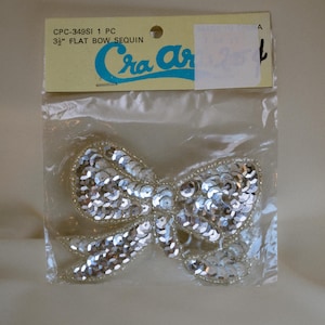 May include: A silver sequin bow, approximately 9 cm wide, in a clear plastic package. The bow is made of many small, shiny sequins and is outlined with small beads. The packaging has the text "CPC-349SI 1 PC" and "Cra Ar".
