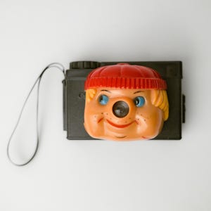 May include: A black plastic film camera featuring a molded orange face with blue eyes and a red beanie cap attached to the front. The camera includes a black wrist strap extending from the side, with the lens positioned as the nose of the face.