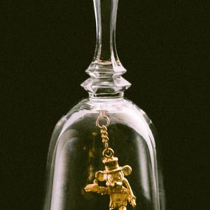 May include: A clear glass bell with a long stem and a gold-coloured charm of a cartoon mouse wearing a hat and holding a tray. The bell is on a black background.