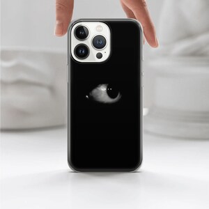 May include: A clear phone case with a black background and a white eye graphic. The phone case is designed for an iPhone with a triple lens camera system.