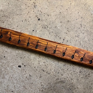 Könnte beinhalten: Eine lange, rechteckige Wandleiste aus einem einzigen Stück naturfarbenem Holz mit warmen Tönen und einer ausgeprägten Maserung. Das Holzbrett ist mit elf gleichmäßig verteilten, kleinen, schwarzen Metallhaken in einer horizontalen Reihe versehen.