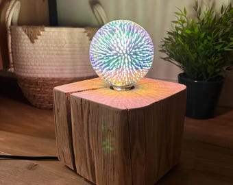 Puede incluir: Lámpara decorativa con una esfera de vidrio 3D que emite luz colorida, colocada sobre una base de bloque de madera natural. La lámpara está enchufada y se encuentra sobre una superficie de madera. Una cesta tejida y una planta en maceta están en el fondo.