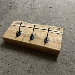 Könnte beinhalten: Ein Holz-Kleiderständer mit drei schwarzen Metallhaken. Der Ständer ist aus recyceltem Holz gefertigt und hat eine natürliche Oberfläche. Die Haken sind einfach und funktional.