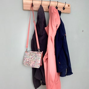Könnte beinhalten: Ein Holz-Kleiderständer mit fünf Haken, an dem eine rosa Jacke, eine dunkelgraue Jacke und eine marineblaue Jacke hängen. Eine kleine gemusterte Handtasche hängt an einem der Haken.