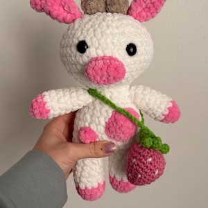 May include: A handmade crochet cow plushie with white and pink accents. The cow has black eyes, pink ears, and a pink nose. It carries a strawberry-shaped bag with a green strap. A cute and cuddly toy.