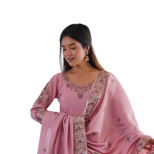 May include: A light pink Anarkali suit featuring long sleeves, a V-neckline, and a flared skirt. The garment is adorned with intricate metallic embroidery and small mirror accents along the hem, neckline, and the matching draped dupatta shawl.