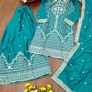 May include: A teal embroidered outfit, comprising a long-sleeved tunic, skirt, and scarf. The tunic and skirt have detailed white floral embroidery on the edges and bodice. The outfit is presented on a wooden surface, with a basket of flowers in the background.