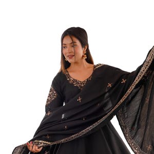 May include: A black Anarkali dress featuring a V-neckline and long sleeves, adorned with intricate copper-toned embroidery and mirror work along the hem and neckline. A matching black dupatta with embroidered borders is draped across the shoulders.
