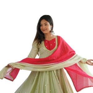May include: A light green Anarkali dress featuring a red bodice with gold embroidery, long sleeves, and a flared skirt adorned with small circular mirrors. A matching light green and hot pink dupatta with gold trim is draped across the front.