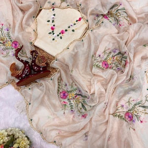 May include: A cream-colored sari with floral embroidery in pink, purple, and yellow. The sari has a scalloped edge with green beads and gold trim. A matching cream-colored top with floral embroidery is also visible.