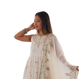 May include: An off-white sleeveless floor-length dress featuring intricate gold embroidery and reflective mirror embellishments on the bodice and skirt. A matching sheer off-white dupatta with a wide gold embroidered border and mirror work is draped.