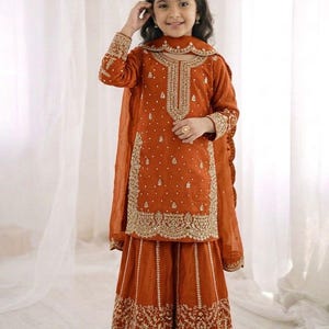 Op de afbeelding: Een roestkleurige, geborduurde salwar kameez set voor een kind. De outfit heeft ingewikkeld gouddraadwerk op de halslijn, mouwen en zoom van de top en palazzo broek. Een bijpassende, doorschijnende dupatta maakt de ensemble compleet, met traditioneel Zuid-Aziatisch design.
