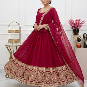 May include: A formal, floor-length burgundy dress with gold embroidery. The dress features a round neckline, long sleeves, and a flowing skirt. Gold embellishments adorn the neckline, cuffs, and hem. A matching sheer scarf drapes over the shoulders.