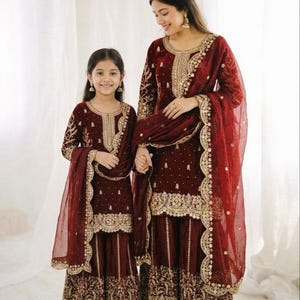 May include: Matching maroon outfits with gold embroidery. The outfits include long-sleeved tops, wide-leg pants, and sheer scarves. The outfits are worn by a woman and a young girl, suggesting a mother-daughter pairing. The outfits are suitable for special occasions.