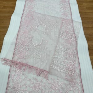 May include: A delicate, light pink embroidered fabric with floral and geometric designs. The fabric has a sheer quality and is adorned with small, sparkling embellishments and fringe. The fabric is displayed on a white surface.