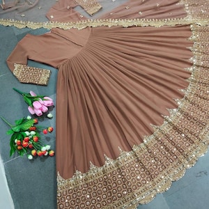 Puede incluir: Un vestido marrón con mangas largas y una falda fluida, adornado con lentejuelas doradas y bordados en el dobladillo. Se incluye una bufanda transparente a juego con ribete dorado y un pequeño bolso. El vestido se muestra sobre una superficie gris con flores.