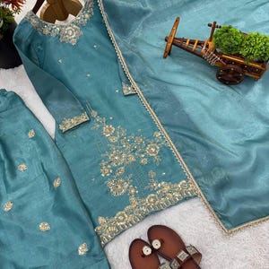 以下が含まれることがあります： ターコイズ色の刺繍が施されたフォーマルな衣装。長袖チュニック、パンツ、お揃いのスカーフが含まれています。チュニックとパンツには金の刺繍とスパンコールが施されています。銀色のアクセントが付いた茶色の革サンダルも写っています。